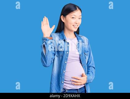 Jeune belle femme chinoise enceinte s'attendant à un bébé qui renonce à dire bonjour heureux et souriant, geste de bienvenue amical Banque D'Images