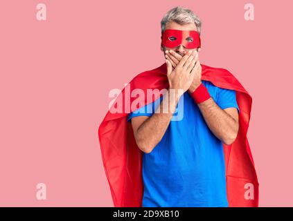 Jeune homme blond portant le super héros crème choqué couvrant la bouche avec les mains pour erreur. Concept secret. Banque D'Images