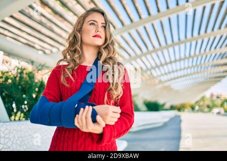 Jeune fille blonde d'expression sérieuse injuriée avec une élingue de bras debout au parc. Banque D'Images