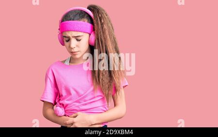 Adorable enfant hispanique fille portant des vêtements de gym et utilisant des écouteurs avec la main sur l'estomac parce que l'indigestion, la maladie douloureuse sentiment malaise. Ache conce Banque D'Images