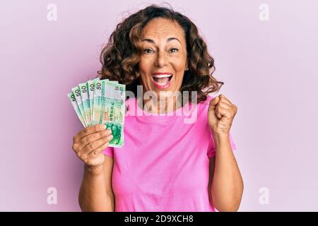 Une femme hispanique d'âge moyen détenant 50 billets de banque en dollars de hong kong criait fière, célébrant la victoire et le succès très enthousiaste avec le bras relevé Banque D'Images