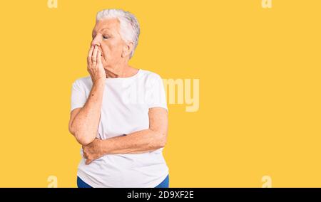Senior belle femme avec les yeux bleus et les cheveux gris portant décontracté blanc Tshirt ennuyé bâilling fatigué couvrant la bouche avec la main. Agité et somnolence. Banque D'Images