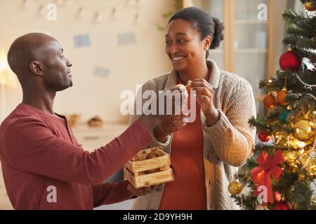 Taille vers le haut portrait d'un couple afro-américain aimant décorant l'arbre de Noël ensemble tout en profitant de la saison des vacances à la maison Banque D'Images