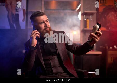 jeune homme en costume, barbier tient la tondeuse dans les mains et parle au téléphone, au travail. dans une pièce sombre et fumée Banque D'Images