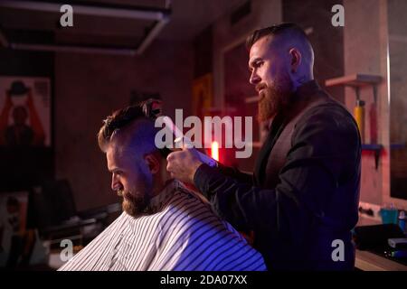 coiffeur homme confiant coupe les cheveux à l'homme élégant et élégant assis dans le salon, les jeunes hommes apprécient le processus de coupe Banque D'Images