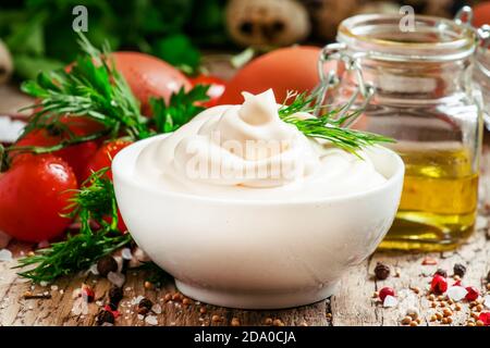 Sauce mayonnaise maison dans un bol blanc, bol avec huile d'olive, œufs, sel, épices, moutarde, herbes, tomates cerises sur fond de bois ancien, sélective Banque D'Images