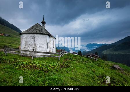 Chapelle blanche au toit de bardeaux sur les prairies Ackernalm alp avec ciel couvert et panorama sur les montagnes du Kaiser, Tyrol, Autriche Banque D'Images