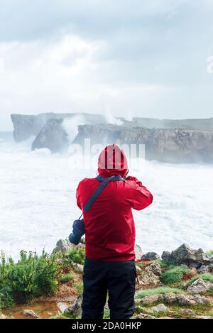 Vue arrière du voyageur anonyme en imperméable prenant en photo mer orageux sur une falaise rocheuse le jour nuageux à l'intérieur Bufones de Llames Pría en Espagne Banque D'Images