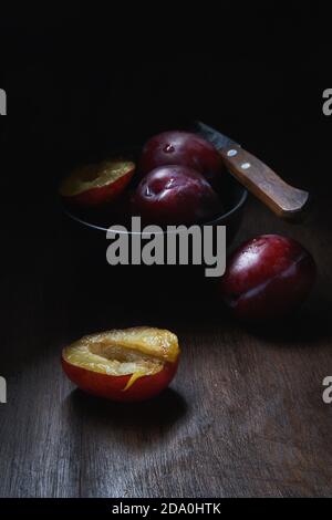 Composition de vie fixe avec bol avec prunes aromatiques juteuses mûres avec gouttes d'eau disposées sur une table en bois avec couteau et fruits coupés en deux Banque D'Images