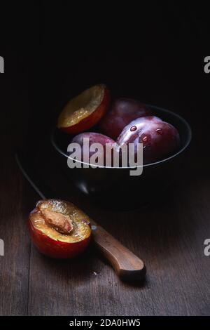 Composition de vie fixe avec bol avec prunes aromatiques juteuses mûres avec gouttes d'eau disposées sur une table en bois avec couteau et fruits coupés en deux Banque D'Images