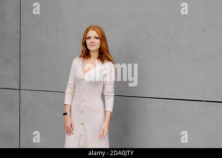 Positive jeune femme avec de longs cheveux rouges portant une robe décontractée et bracelet intelligent regardant la caméra tout en se tenant contre le béton mur Banque D'Images