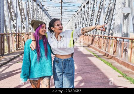 Jeunes filles à la mode avec bras et jambes tatoués debout pont et pointant et regardant loin Banque D'Images