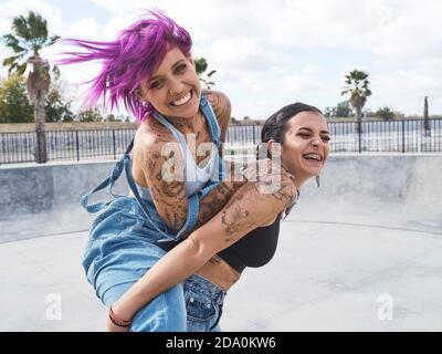 Femme ravie avec les cheveux roses piggydoring amie joyeuse avec tatouages tout en s'amusant ensemble le week-end Banque D'Images