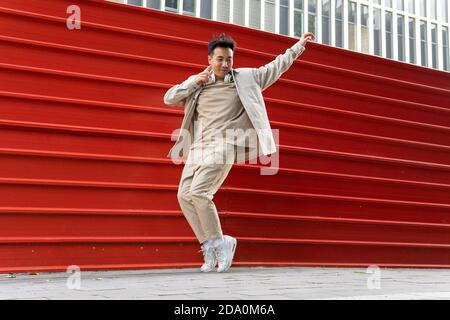 Homme asiatique plein de corps joyeux en tenue décontractée à l'écoute chanson préférée dans un casque sans fil tout en dansant le long du métal rouge clôture Banque D'Images