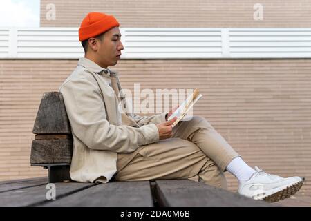 Vue latérale asiatique homme portant des vêtements décontractés lisant fascinant réservez en vous asseyant sur un banc en bois dans la rue Banque D'Images