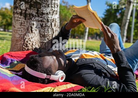 Homme ethnique calme dans un casque allongé sur une couverture et la lecture un roman intéressant tout en se relaxant le jour d'été dans le parc Banque D'Images