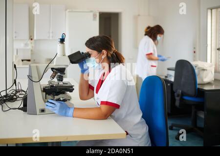 Femmes technologues en uniformes et masques stériles travaillant avec microscope en laboratoire Banque D'Images