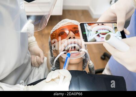 Du dessus de la récolte, dentiste méconnaissable prenant une photo des dents de patient sur smartphone tout en injectant au patient se préparant pour traitement dentaire Banque D'Images