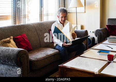 Un homme joyeux et agé examine son plan d'affaires tout en se reposant canapé confortable et doux près de la table avec des papiers Banque D'Images