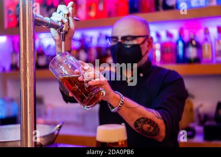 Vue latérale d'un barkeeper sérieux dans des lunettes de vue qui versent de la bière verre pendant qu'il travaille en bar pendant une pandémie de coronavirus Banque D'Images