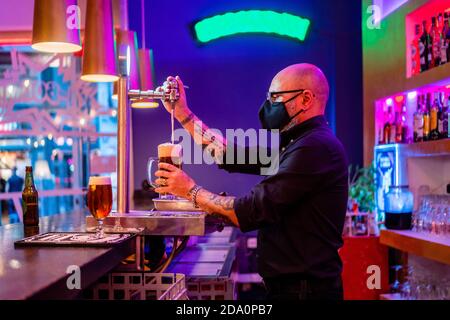 Vue latérale d'un barkeeper sérieux dans des lunettes de vue qui versent de la bière verre pendant qu'il travaille en bar pendant une pandémie de coronavirus Banque D'Images