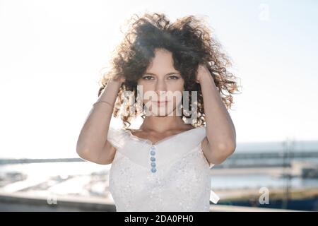 Jeune femme tendre portant dentelle robe de mariée touchant la tête et regarder l'appareil photo sur un arrière-plan flou de la ville Banque D'Images