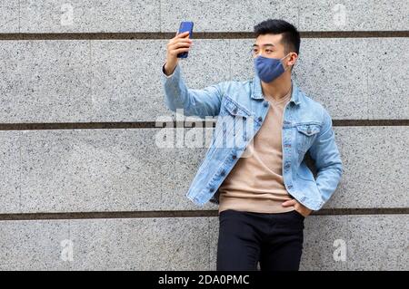 Homme asiatique portant un masque médical de protection debout près du bâtiment et prise de selfie sur smartphone pendant l'épidémie de coronavirus en ville Banque D'Images