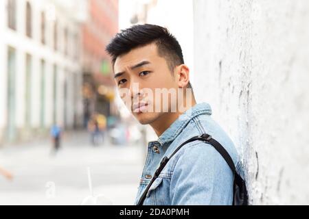 Vue latérale d'un jeune homme asiatique sérieux étudiant en denim veste avec perçage dans l'oreille debout près du mur de la ville construire et regarder la caméra Banque D'Images