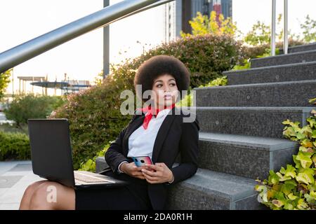 Entrepreneur afro-américain sérieux avec cheveux afro travaillant sur ordinateur portable asseyez-vous dans les escaliers en regardant loin Banque D'Images