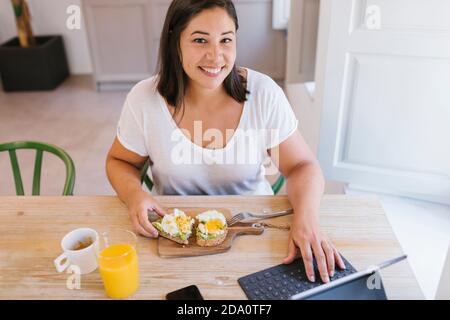 Bonne femme souriante ayant un petit déjeuner sain et travaillant à la maison avec une tablette Banque D'Images