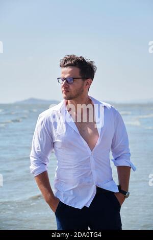 Jeune homme élégant en vêtements et lunettes tendance avec les mains dans les poches derrière la mer ondulée sous le ciel Banque D'Images