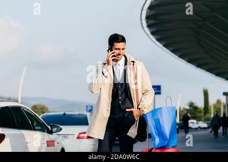 Positif homme avec des cheveux noirs dans des vêtements formels debout avec sacs de shopping et parler au téléphone contre un bâtiment moderne Banque D'Images