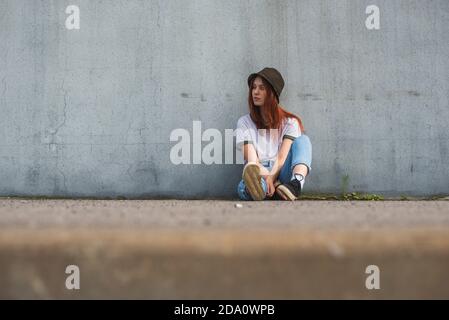 Tout le corps de l'adolescence tendance rouge cheveux femelle élève dans tenue décontractée et chapeau incliné vers l'arrière sur béton gris mur sur la rue urbaine Banque D'Images