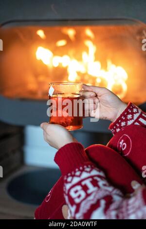 Vue latérale d'une femme méconnue dans un chandail chaud rouge assis Avec une tasse de thé près de la cheminée et profiter de Noël confortable nuit Banque D'Images