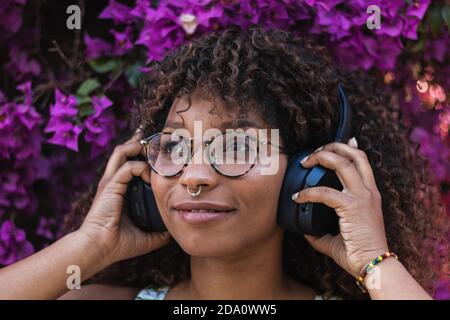 Femme afro-américaine avec piercing et lunettes souriantes et à l'écoute pour écouter de la musique dans un casque contre les broussailles Banque D'Images