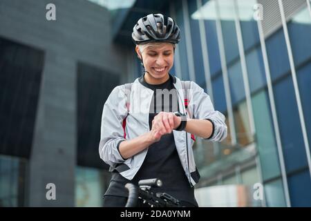 Une jeune cycliste regarde la montre-bracelet sur le fond d'un bâtiment moderne le jour d'été dans la ville Banque D'Images