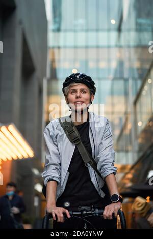 Femme dans un casque sur une bicyclette près d'un grand bâtiment avec des fenêtres en verre, gros plan Banque D'Images