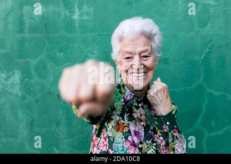 Gaie vieille femme grise aux cheveux dans élégant chemisier coloré ayant amusez-vous et boxe à l'appareil photo tout en vous tenant contre le mur vert Banque D'Images