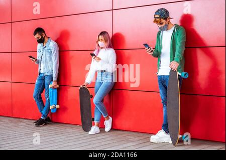 Adolescents avec masque pour le visage messages texte de téléphone mobile s'appuyant sur Mur urbain dans la distanciation sociale - Groupe d'amis multiraciaux utilisation du cellulaire Banque D'Images