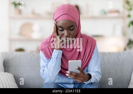 Femme musulmane noire préoccupée assise avec un smartphone entre les mains Accueil Banque D'Images