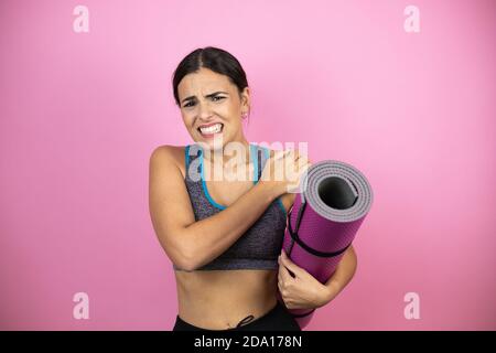 Jeune belle femme portant des vêtements de sport sur un arrière-plan isolé rose tenue une attelle dans sa main avec une douleur sur son épaule et une expresssi douloureuse Banque D'Images