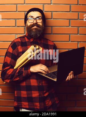 Homme barbu, longue barbe. Brutal caucasien sourire heureux non rashen taille basse tenant un ordinateur portable et des rouleaux de papier d'artisanat en rouge noir damier chemise avec chapeau et des lunettes sur fond de mur de briques brunes studio Banque D'Images