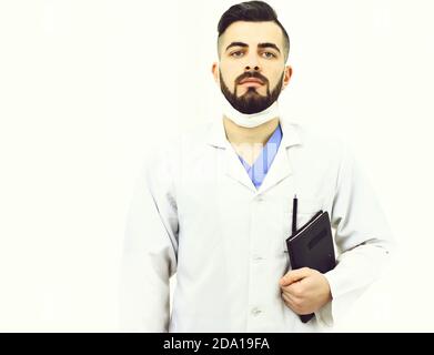 Le médecin avec la barbe tient un carnet avec des antécédents de maladie actuelle. Concept de traitement et services d'ambulance. Homme au visage confiant, sous un manteau médical blanc. Chirurgien dans le masque isolé sur fond blanc Banque D'Images