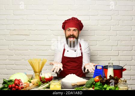 Cuisinez avec le visage heureux en uniforme, assis près d'une table de cuisine avec des légumes, des pâtes et des ustensiles de cuisine. Le chef prépare le repas. Homme avec la barbe travaille avec la farine sur fond de brique blanche. Concept du processus de cuisson. Banque D'Images