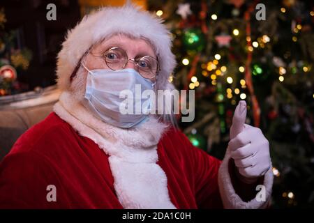 Le Père Noël en masque médical. Portrait positif Santa montrant le pouce vers le haut assis près de l'arbre de Noël. Bonne année Covid 2021 Banque D'Images
