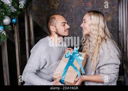 Le jeune couple aimant échange des cadeaux pour la nouvelle année. Le mari embrasse sa femme et donne un cadeau pour la nouvelle année. Cadeau de Noël et du nouvel an à la maison. Banque D'Images