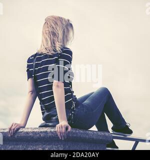 Vue arrière d'une jeune femme de mode assise contre un ciel nuageux. Modèle féminin élégant en t-shirt à col roulé extérieur Banque D'Images