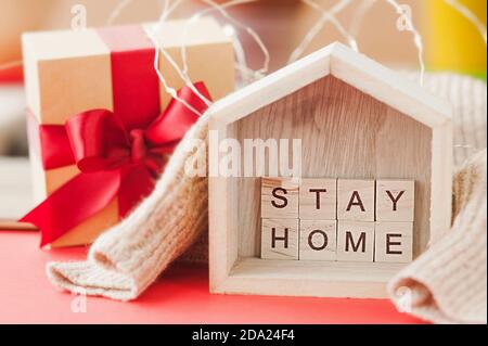 restez à la maison en lettres carrées dans une petite maison en bois sur pull beige, guirlandes brillantes et boîte cadeau avec ruban rouge. avertissement pour th Banque D'Images