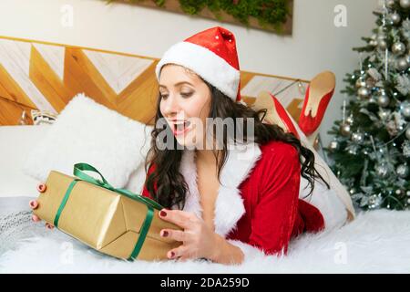 Jolie fille souriant avec cadeau près de l'arbre de Noël. Jeune fille en vêtements et chapeau du Père Noël Banque D'Images