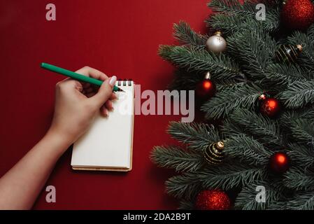 Une femme écrit une liste de shopping ou de tâches dans un carnet sur un fond rouge à côté des branches et des décorations du nouvel an. Maquette. Banque D'Images